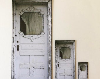 Könnte beinhalten: Drei gerahmte Drucke einer weißen Tür mit einem dekorativen Fenstersprossenmuster. Die Tür ist verwittert und hat einen abgenutzten Look. Die Drucke sind in verschiedenen Größen, wobei der größte Druck in der Mitte ist.