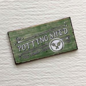 May include: A green wooden sign with the text "Potting Shed" and the words "Pots, Plants, Seeds" underneath. The sign has a rustic, distressed look and features a small circle with a gardening fork and trowel.