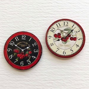 Op de afbeelding: Twee ronde rood-witte klokken met zwarte Romeinse cijfers voor de uren en zwarte wijzers. De wijzerplaten van de klokken tonen een kersenboomtak met rode kersen en groene bladeren. Een wijzerplaat heeft de tekst "Joseph Tyler Co. Boston" erop. De andere wijzerplaat heeft de tekst "Cerise Commune" erop.