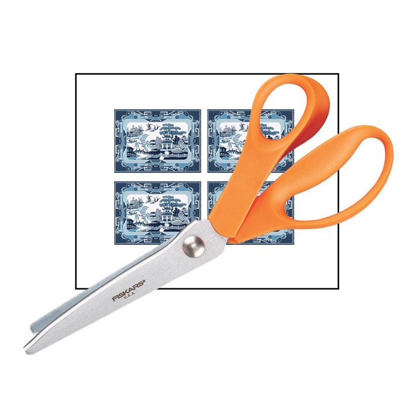 May include: Scissors with orange handles and a silver blade, featuring the text "FISKARS&reg; USA" on the blade. The scissors are positioned on top of a sheet of paper with four blue and white patterned squares.