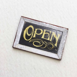 Puede incluir: Un letrero de madera de estilo vintage con marco blanco y fondo negro. La palabra "ABIERTO" est&aacute; escrita en letras doradas con remolinos decorativos.