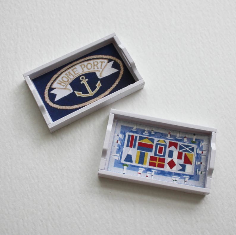 May include: Two white wooden trays with nautical designs. The first tray has a blue background with a white rope border and an anchor with the text "Home Port". The second tray has a blue and white background with a pattern of colorful nautical flags.