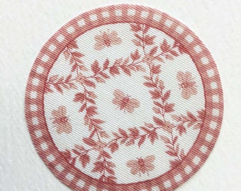 Puede incluir: Un mantel individual redondo de tela rosa y blanca con un diseño floral. El mantel individual tiene un borde rosa con un patrón de cuadros blancos.