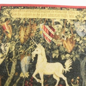 May include: A tapestry featuring a white unicorn with a red, white, and blue flag in the background. The tapestry has a quote in the top border: "There are amongst certain beasts of the round table born to serve the unicorn on the quest whatever might betide."