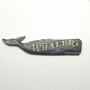 May include: A metal whale-shaped sign with the text "THE WHALERS" in gold lettering. The sign is dark gray with a textured surface and a whale tail on the left side. The whale's body has a detailed design.