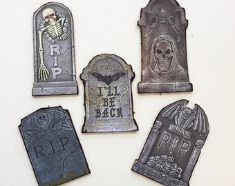 May include: Five flat tombstone-shaped decorations featuring various designs: a skeleton with red eyes and the text R.I.P., a hooded skull, a bat with the text I'LL BE BACK, a winged skull with R.I.P., and a gargoyle with R.I.P. and skulls.