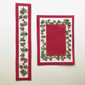 May include: Two red fabric pieces with a white border and a green holly leaf and red berry design. The larger piece has a red center with a white border and the smaller piece is a long strip.