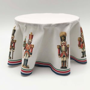May include: White round tablecloth with a red, green and blue border. The tablecloth features a repeating pattern of nutcracker soldiers in red and blue uniforms.