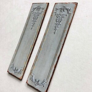 May include: Two decorative metal wall panels with a distressed, aged, light blue finish. Each panel features a raised, ornate design in the center and a brown border.