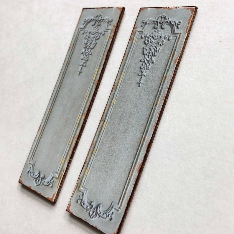 May include: Two decorative metal wall panels with a distressed, aged, light blue finish. Each panel features a raised, ornate design in the center and a brown border.