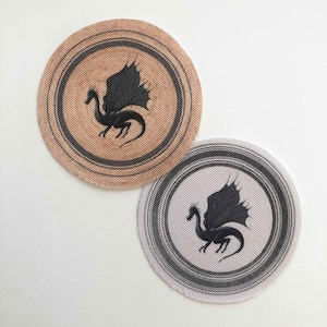 以下が含まれることがあります： 翼を広げた黒いシルエットのドラゴンが描かれた、丸い布製のコースターが2つ。1つのコースターは同心円状の茶色で、もう1つは同心円状のグレーの白です。