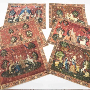 May include: Six square tapestry placemats featuring a medieval scene with a unicorn, a lion, and a woman in a forest. The placemats have a red background with gold trim.