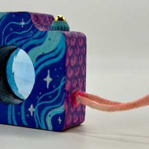 May include: A wooden camera ornament painted with a celestial design in shades of blue, purple, and teal. The camera features a moon-shaped lens and a pink cord. The ornament has a gold-colored button on top.