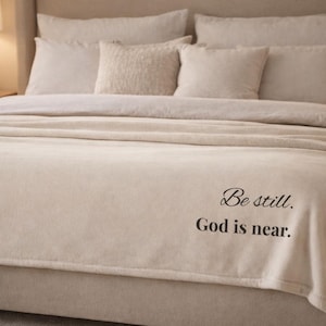 May include: A beige bed with a plush blanket and pillows. The blanket has the text "Be still. God is near." in black script. The bed is made up with several pillows and a soft blanket.