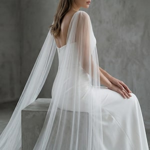May include: A white wedding dress with a flowing, sheer cape. The dress has a simple, elegant design with a fitted bodice and a long, flowing skirt. The cape drapes over the shoulders and extends to the floor, creating a dramatic effect.
