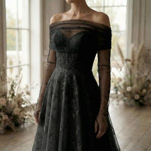 May include: A black off-the-shoulder gown with a floral pattern. The dress features sheer, long sleeves and a bodice with delicate bead embellishments. The skirt flows gracefully, creating an elegant silhouette, perfect for formal events.