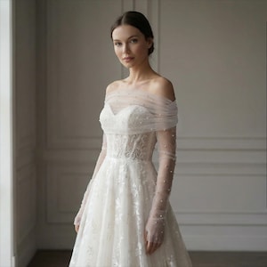 May include: An off-white wedding dress with a sweetheart neckline and sheer, long sleeves adorned with small pearls. The dress features a fitted bodice with floral lace detailing and a flowing skirt. The model is wearing the dress in a well-lit room.