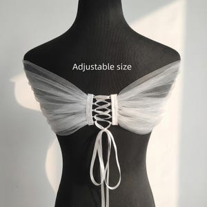 May include: A white tulle bridal capelet with a corset-style back. The capelet is displayed on a black mannequin. The text "Adjustable size" is printed on the image. The capelet has a delicate, airy appearance, suitable for weddings or special occasions.