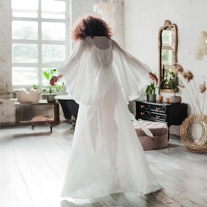 K&ouml;nnte beinhalten: Ein flie&szlig;endes wei&szlig;es Kleid mit transparenten, kapuzenartigen &Auml;rmeln. Das Kleid hat ein eng anliegendes Oberteil und einen langen, flie&szlig;enden Rock. Die Person befindet sich in einem hellen Raum mit einem gro&szlig;en Fenster und einem Spiegel im Vintage-Stil.