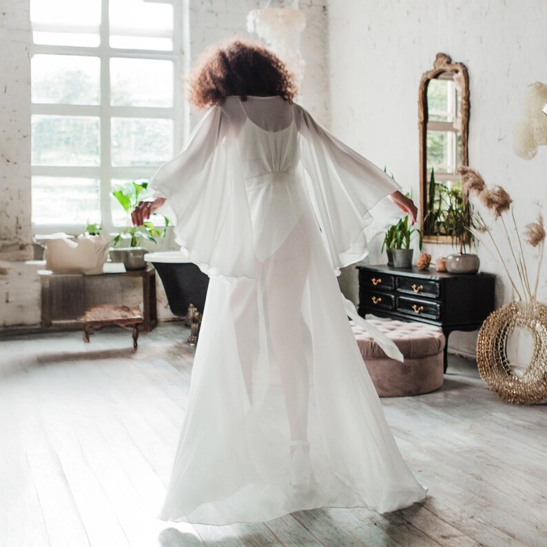 K&ouml;nnte beinhalten: Ein flie&szlig;endes wei&szlig;es Kleid mit transparenten, kapuzenartigen &Auml;rmeln. Das Kleid hat ein eng anliegendes Oberteil und einen langen, flie&szlig;enden Rock. Die Person befindet sich in einem hellen Raum mit einem gro&szlig;en Fenster und einem Spiegel im Vintage-Stil.
