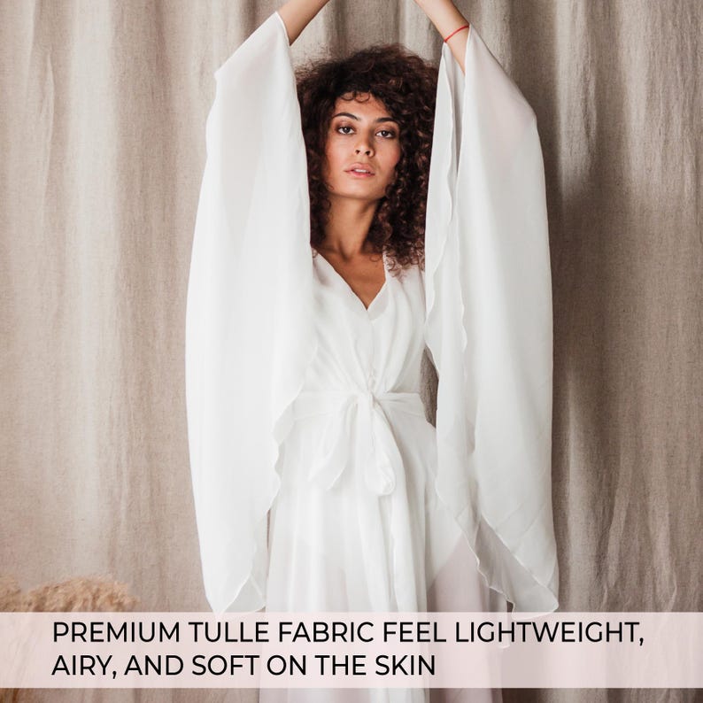 May include: A white tulle fabric dress with long, flowing sleeves and a V-neckline. The dress is tied at the waist with a matching fabric belt. The text at the bottom of the image reads: PREMIUM TULLE FABRIC FEEL LIGHTWEIGHT, AIRY, AND SOFT ON THE SKIN.