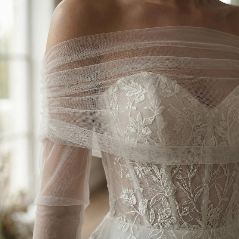 May include: Off-the-shoulder wedding dress with a sweetheart neckline. The bodice features intricate floral lace detailing. The sheer, layered fabric drapes over the shoulders and arms, creating an elegant, romantic look.