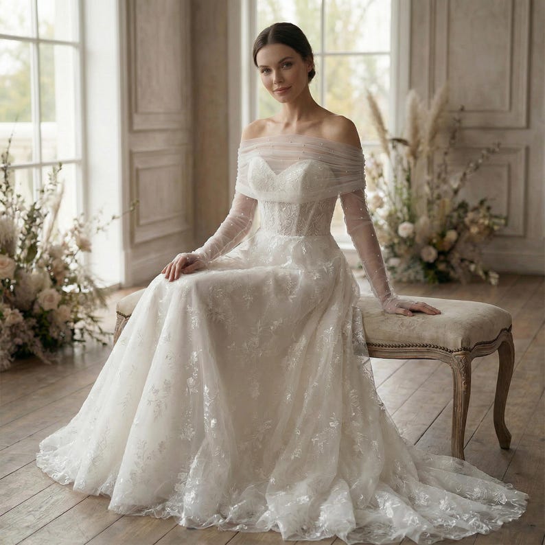 May include: A white wedding dress with an off-the-shoulder neckline and long, sheer sleeves. The dress features intricate floral lace detailing and a flowing skirt. The bride is seated on a cushioned bench in a well-lit room.