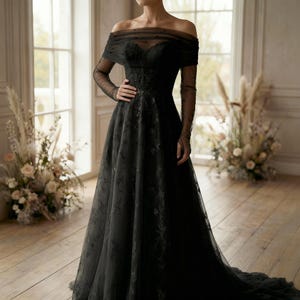May include: A black off-the-shoulder gown with long, sheer sleeves. The dress features a fitted bodice with floral detailing and a flowing skirt. The gown is floor-length and has a train. The background includes windows and floral arrangements.