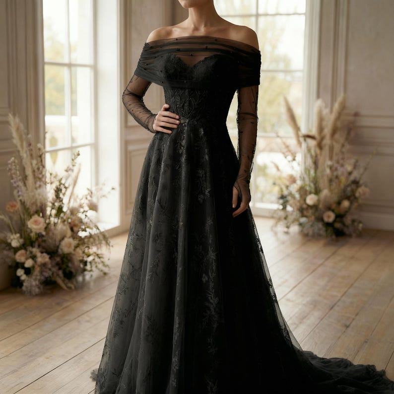 May include: A black off-the-shoulder gown with long, sheer sleeves. The dress features a fitted bodice with floral detailing and a flowing skirt. The gown is floor-length and has a train. The background includes windows and floral arrangements.