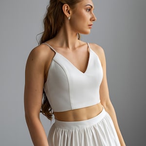 May include: A white crop top with a V-neck and thin straps, paired with a matching white skirt. The outfit is a bridal or formal wear ensemble, suitable for a wedding or special event.