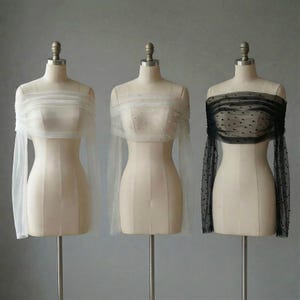 May include: Three sheer, off-the-shoulder tops on dress forms. One is white, one is white with pearl accents, and the third is black with small, dark embellishments. Each has long sleeves and a gathered neckline.