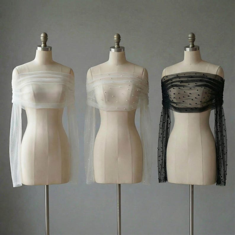May include: Three sheer, off-the-shoulder tops on dress forms. One is white, one is white with pearl accents, and the third is black with small, dark embellishments. Each has long sleeves and a gathered neckline.