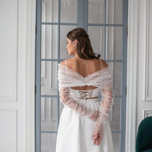 May include: A white bridal ensemble featuring a corset-style top with a lace-up back and a flowing skirt. A sheer, long-sleeved wrap adorned with pearls is draped over the shoulders. The outfit is designed for a wedding or special occasion.