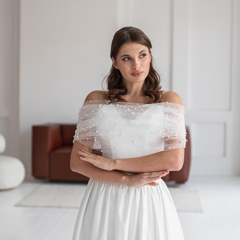 May include: A white wedding dress with a tulle overlay adorned with small, round pearls. The dress features a fitted bodice and a flowing skirt. The tulle overlay is draped over the shoulders, creating a delicate, elegant look. The dress is suitable for a bride.