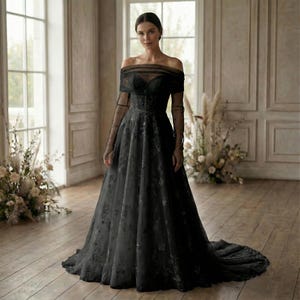 May include: A black off-the-shoulder wedding gown with long sleeves. The dress features a floral pattern on the skirt and bodice, and a sheer overlay on the sleeves. The gown has a long train and is displayed in a well-lit room.