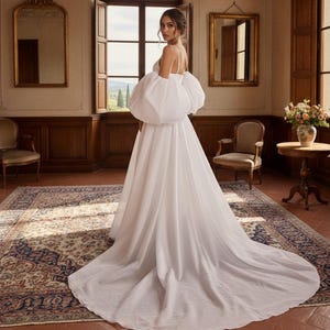 May include: A white wedding dress with a flowing skirt and dramatic, oversized puff sleeves. The dress features spaghetti straps and a long train. The image is set in a room with a large window and antique furniture, suggesting a classic wedding theme.