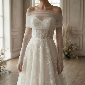 May include: Off-the-shoulder ivory wedding dress with floral lace detailing. The gown features a sweetheart neckline, fitted bodice, and a flowing skirt. Sheer, long sleeves and a draped overlay add an elegant touch to this bridal design.