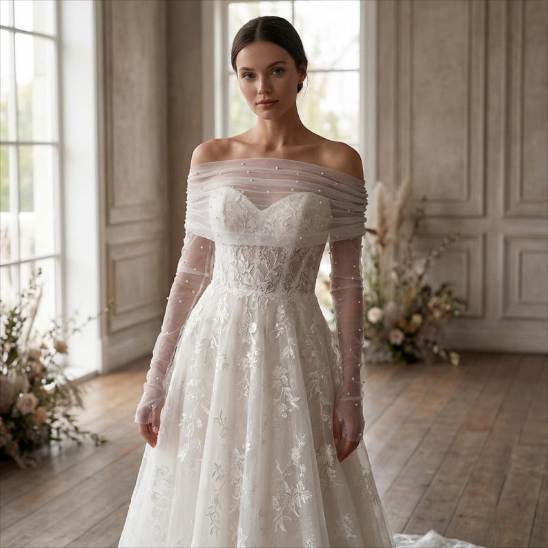 May include: A white off-the-shoulder wedding dress with long, sheer sleeves. The bodice features intricate lace detailing, and the neckline is adorned with small pearls. The dress has a full skirt and a flowing train, perfect for a bride.