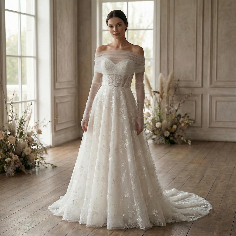 May include: A white wedding dress with an off-the-shoulder neckline and long sleeves. The dress features a fitted bodice with floral lace detailing and a flowing skirt. The gown has a long train and is made of a sheer, lightweight fabric.