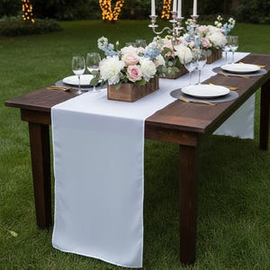 May include: A long, dark wooden table set for an outdoor event. A white table runner is centered on the table, with floral arrangements in wooden boxes, plates, glasses, and gold-colored cutlery. The flowers are white, pink, and blue.