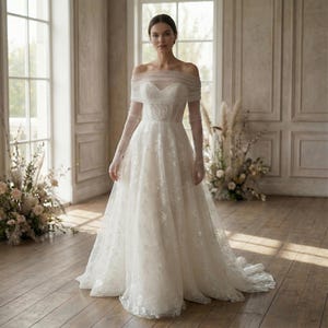 May include: A white off-the-shoulder wedding dress with long sleeves. The gown features a sweetheart neckline and a full skirt with floral lace detailing. The model is standing in a well-lit room with large windows and floral arrangements.