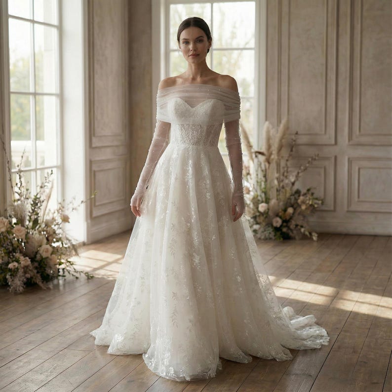 May include: A white off-the-shoulder wedding dress with long sleeves. The gown features a sweetheart neckline and a full skirt with floral lace detailing. The model is standing in a well-lit room with large windows and floral arrangements.