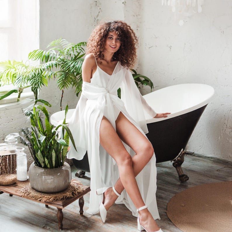 May include: A white, flowing robe with a wrap-around design and long sleeves. The robe is paired with white heeled sandals. The setting includes a black and white bathtub and potted plants, creating a spa-like atmosphere.