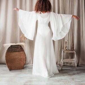 K&ouml;nnte beinhalten: Ein wei&szlig;es, flie&szlig;endes Kleid mit weiten Glocken&auml;rmeln. Das Kleid hat ein eng anliegendes Oberteil und einen langen, durchsichtigen Rock. Das Model ist dem Betrachter abgewandt und hat die Arme ausgestreckt. Ein geflochtener Korb und ein kleiner Holzschemel sind im Hintergrund.