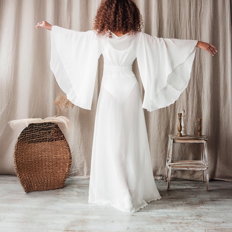 K&ouml;nnte beinhalten: Ein wei&szlig;es, flie&szlig;endes Kleid mit weiten Glocken&auml;rmeln. Das Kleid hat ein eng anliegendes Oberteil und einen langen, durchsichtigen Rock. Das Model ist dem Betrachter abgewandt und hat die Arme ausgestreckt. Ein geflochtener Korb und ein kleiner Holzschemel sind im Hintergrund.