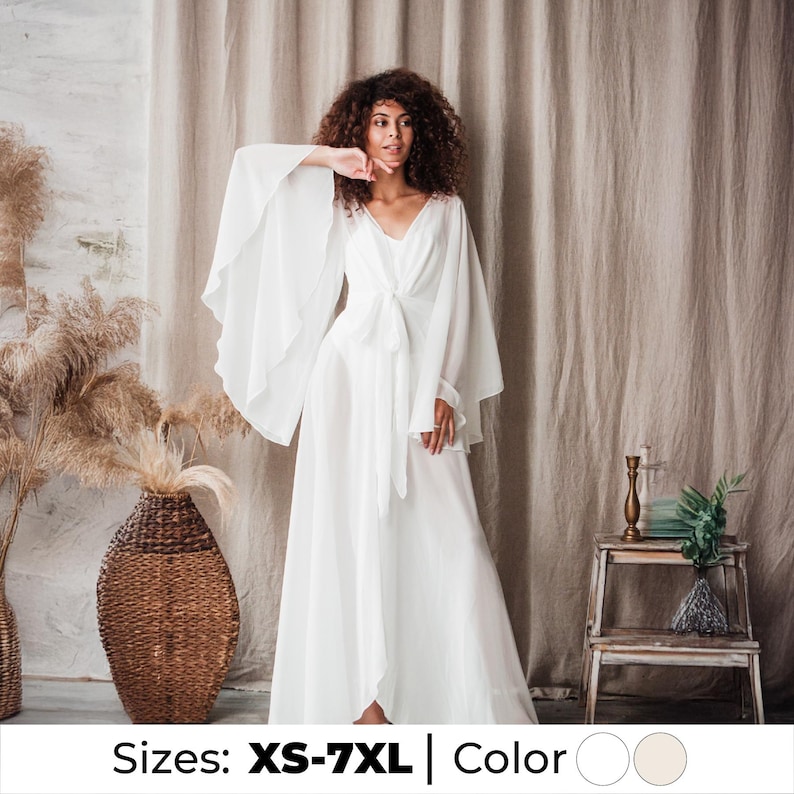 May include: A white, flowing dress with wide sleeves and a V-neckline. The dress is tied at the waist with a matching fabric belt. The model is wearing the dress in front of a neutral backdrop. Sizes available: XS-7XL.