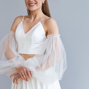 May include: A white bridal top with a V-neckline and spaghetti straps. The top features sheer, ruffled sleeves. The model is wearing a matching white skirt. The outfit is perfect for a wedding or special occasion.
