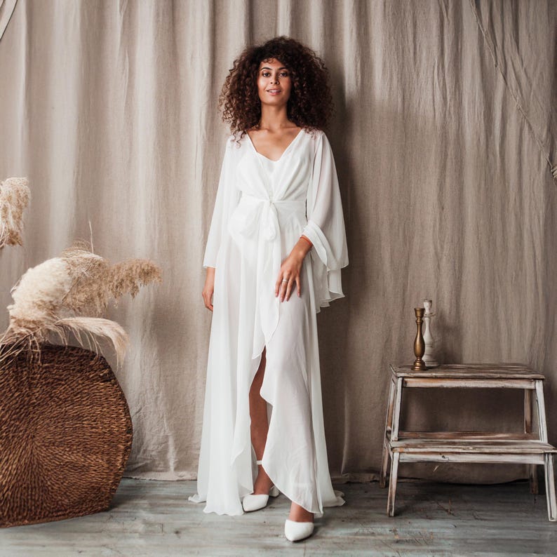 K&ouml;nnte beinhalten: Ein langes, wei&szlig;es Kleid mit weiten &Auml;rmeln und einem V-Ausschnitt. Das Kleid hat eine Schleife in der Taille und einen hohen Schlitz vorne. Das Model tr&auml;gt wei&szlig;e Schuhe. Der Hintergrund ist ein neutrales Beige.