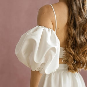 Könnte beinhalten: Weißes Kleid mit Puffärmel und Spaghettiträgern. Das Kleid hat ein figurbetontes Oberteil und einen Rock. Der Ärmel ist an Schulter und Handgelenk gerafft. Der Hintergrund ist einfarbig rosa.