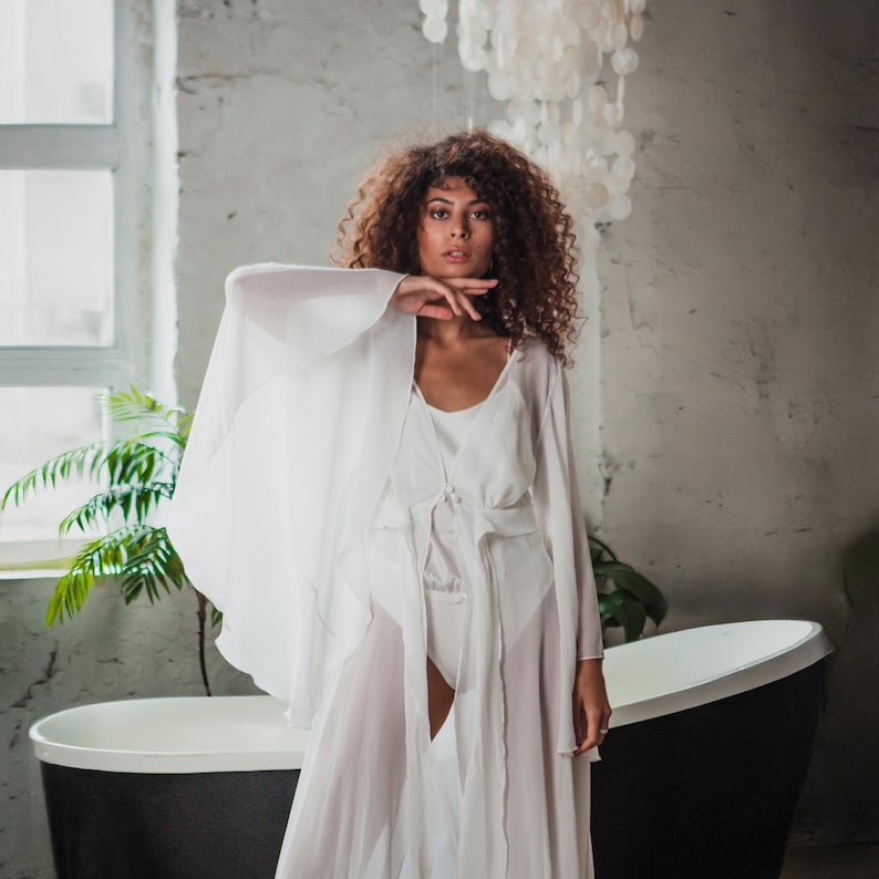 May include: A white sheer robe and matching lingerie set. The robe has long, flowing sleeves and a tie waist. The lingerie set includes a camisole and shorts. The background includes a black bathtub and a window.